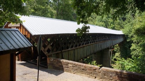 A covered wooden bridge with a metal roof, surrounded by lush greenery and trees.