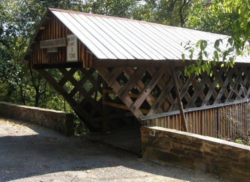 A wooden covered bridge with a metal roof, surrounded by trees and a gravel path. A weight limit sign is visible.