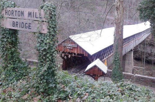 A covered bridge surrounded by trees and greenery, with a sign reading "Horton Mills Bridge" nearby.