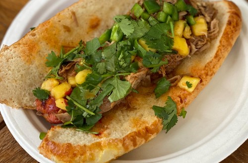 A sandwich filled with pulled meat, mango salsa, green peppers, and cilantro on a toasted bun, served on a paper plate.