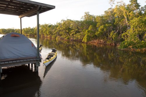 A tent on a dock beside calm water, with kayaks tied up and lush greenery lining the shore.