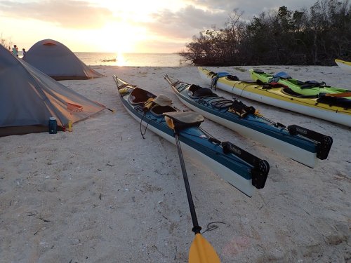 Kayaks on a sandy beach at sunset, with tents and trees in the background.