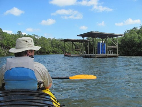 A person in a kayak paddles towards a dock with a blue structure, surrounded by lush greenery and a clear sky.