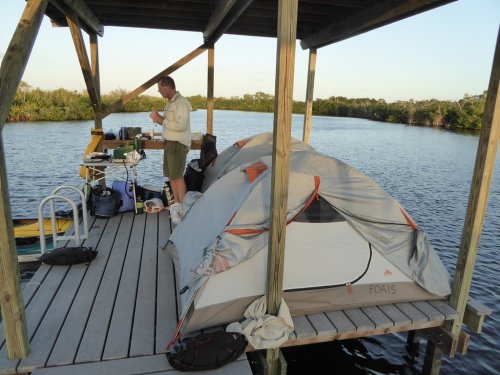 A person prepares food on a dock beside a tent, with water and trees in the background during sunset.