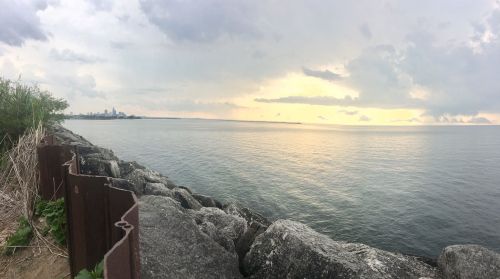 A serene lakeside view with rocky shoreline, calm water, and a cloudy sky at sunset, featuring a distant city skyline.