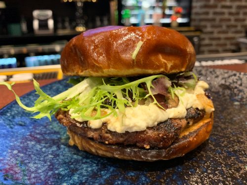 A close-up of a juicy burger on a brioche bun, topped with lettuce and creamy sauce, served on a blue plate.