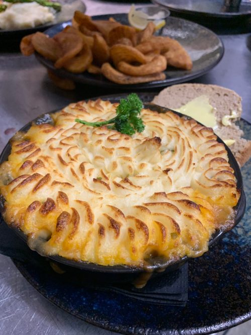 A serving of shepherd's pie with a golden, crispy topping, garnished with parsley, alongside bread and fried snacks.