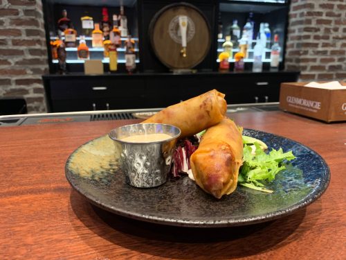 A plate with two spring rolls, a small cup of dipping sauce, and a side of mixed greens on a wooden bar.