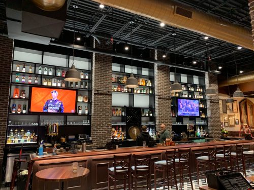 A stylish bar with a brick wall, shelves of liquor, and two TVs displaying sports, with bar stools in front.