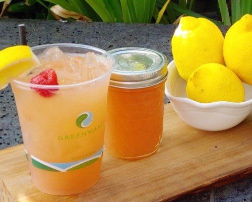 A refreshing drink with a lemon slice and raspberry, alongside a jar of lemon syrup and fresh lemons on a wooden tray.