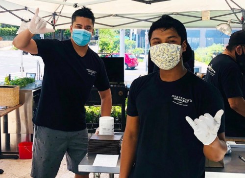 Two masked individuals in black shirts give a thumbs-up and shaka sign at a food stall under a tent.