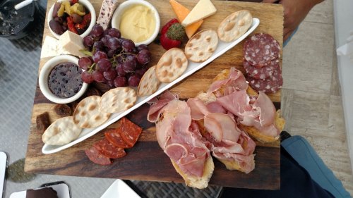 A wooden platter featuring assorted meats, cheeses, crackers, grapes, olives, and a small bowl of jam.