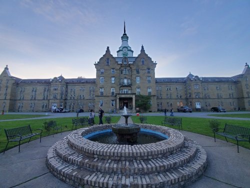 Historic building with a clock tower, surrounded by a fountain and green lawn, at dusk with people nearby.