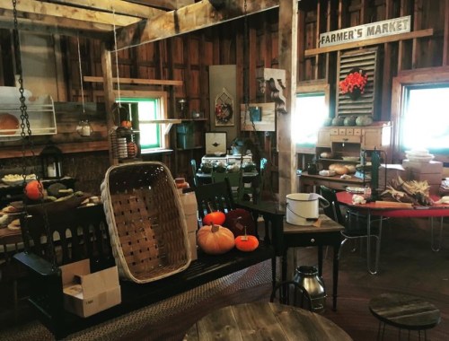 A rustic barn interior featuring a farmer's market setup with produce, baskets, and wooden furniture.