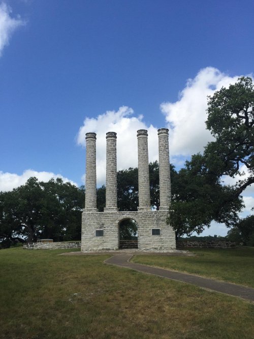 Four tall stone chimneys stand against a blue sky with clouds, surrounded by green grass and trees.