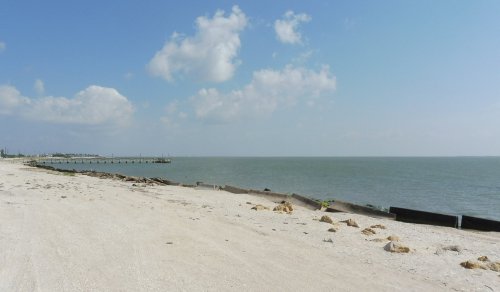 A sandy beach with a calm sea, dotted with clouds in the sky and a distant pier along the shoreline.