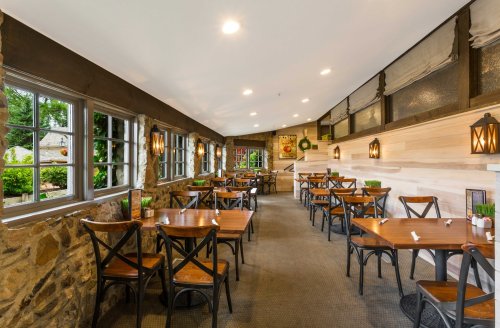 Cozy restaurant interior with wooden tables, stone walls, and large windows overlooking greenery. Warm lighting enhances ambiance.