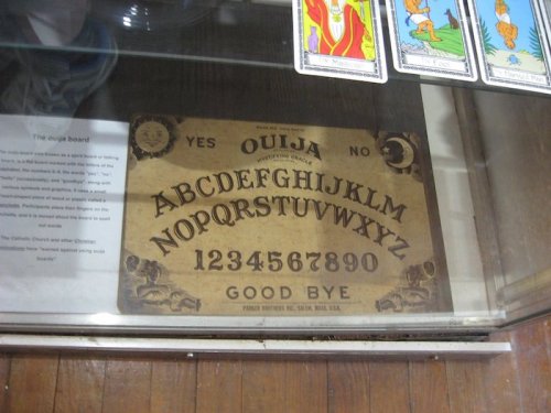 An ouija board displayed in a glass case, featuring letters, numbers, and the words "YES," "NO," and "GOOD BYE."