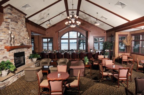 Cozy restaurant interior with a stone fireplace, bar, and tables overlooking a lake through large windows.