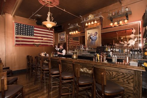 A vintage bar with wooden stools, an American flag, and a bartender behind the counter. Jazz decor is visible.