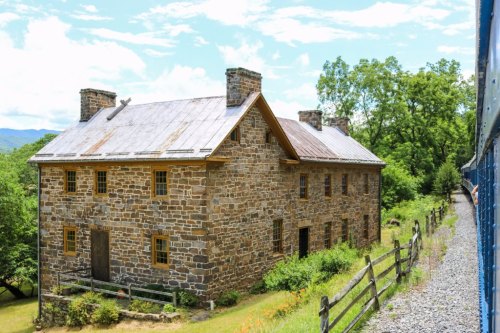 A historic stone building with a metal roof, surrounded by greenery and a wooden fence, under a blue sky.