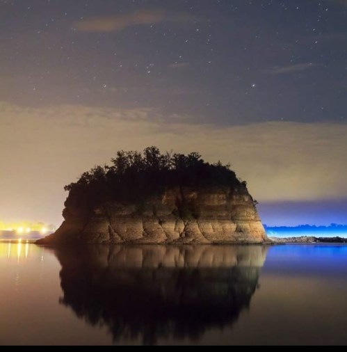 A serene night scene of a rocky island reflecting in calm water, under a starry sky with distant city lights.