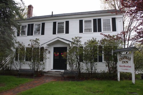 A charming white bed and breakfast with black shutters, surrounded by greenery and featuring a welcoming sign.