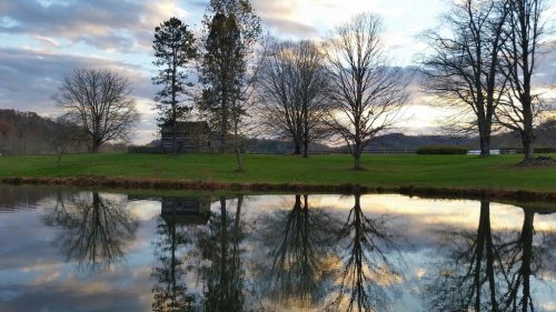 A serene landscape featuring trees and a cabin reflected in calm water under a cloudy sky at sunset.