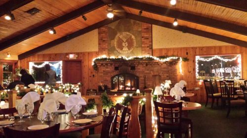 Cozy restaurant interior with wooden beams, a fireplace, and festive lights, set for dining with white tablecloths.