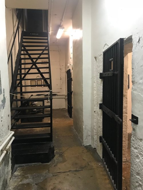 A dimly lit corridor with a metal staircase and two barred doors leading to cells.