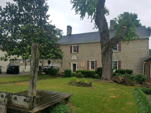 Historic stone building with a gray roof, surrounded by greenery and a wooden structure in the foreground.