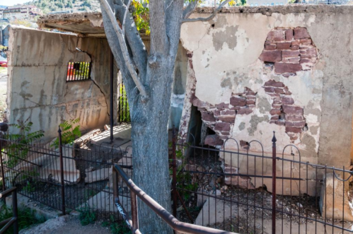 Abandoned building with crumbling walls, a tree growing nearby, and a rusty fence surrounding the area.