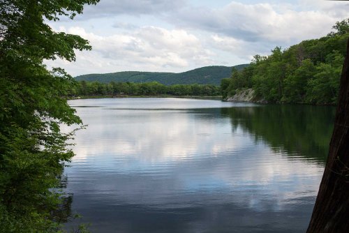 A serene lake surrounded by lush greenery and rolling hills under a cloudy sky. Reflections shimmer on the water's surface.