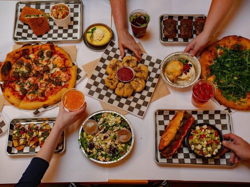 A table spread with various dishes: pizza, salads, cookies, drinks, and desserts, with hands reaching for food.