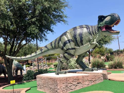 A large, green and black striped dinosaur statue stands on a mini-golf course, surrounded by trees and landscaping.