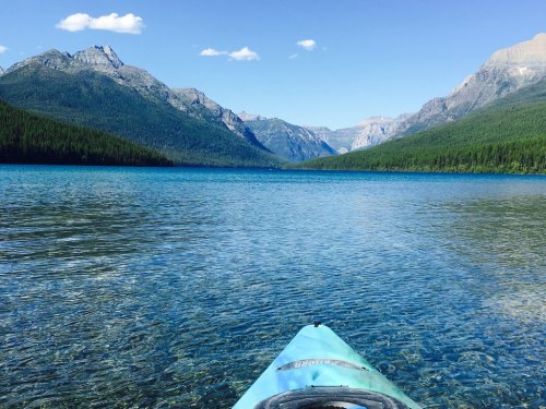 A kayak on clear blue water, surrounded by mountains and lush green forests under a bright blue sky.