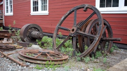 Rusty old wheels and machinery parts resting against a red wooden building, surrounded by greenery.