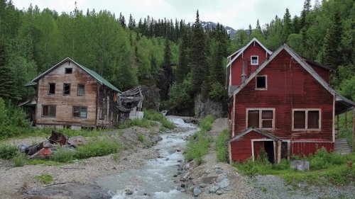 Abandoned wooden houses beside a rocky stream, surrounded by lush green trees and mountains in the background.