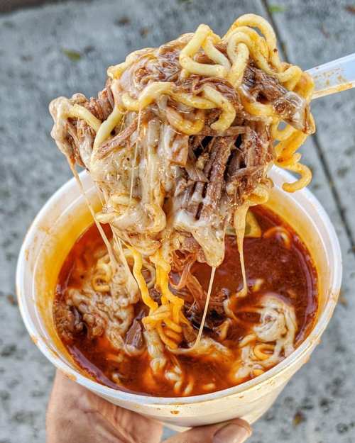 A hand holding a cup of cheesy beef ramen, with noodles and melted cheese stretching from the cup.