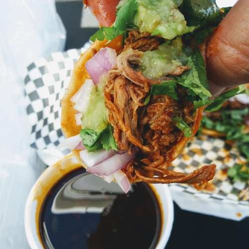 A hand holding a taco filled with shredded meat, onions, and avocado, with a cup of sauce in the background.