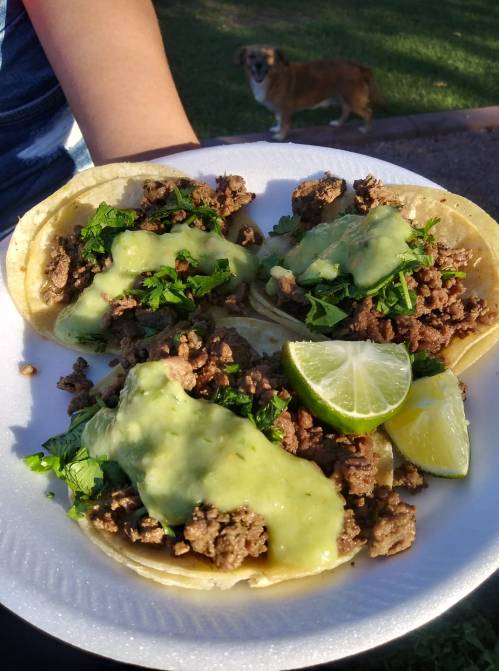 A plate of three tacos filled with seasoned meat, topped with green sauce and cilantro, with lime wedges on the side.