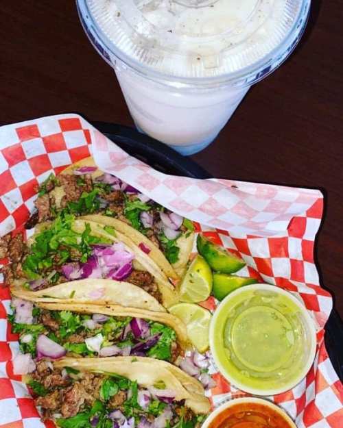 A basket of tacos with beef, onions, and cilantro, served with lime wedges and sauces, alongside a drink.
