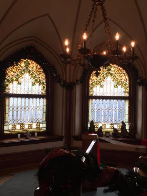 Two ornate stained glass windows adorned with greenery, illuminated by a chandelier in a festive interior setting.