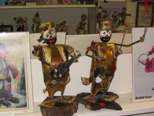 Two whimsical metal sculptures of clowns playing guitar and holding a balloon, displayed on a shelf among other art.