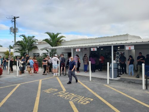 A crowd of people stands in line outside a building with palm trees, while a man walks past a "No Parking" sign.