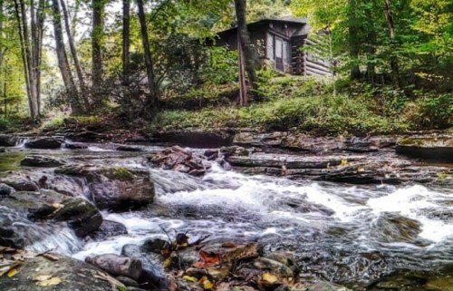 A serene stream flows over rocks, with a log cabin nestled among lush green trees in the background.
