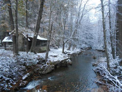 A snowy landscape featuring a rustic cabin by a gently flowing stream, surrounded by trees and winter scenery.