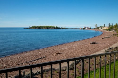 A serene beach scene with calm blue water, sandy shore, and green trees under a clear blue sky.