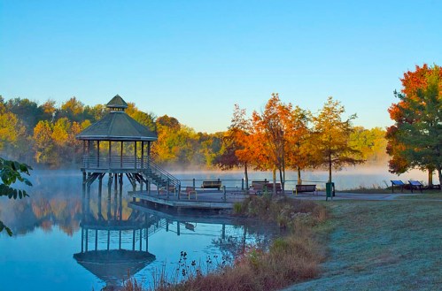 A serene lakeside scene with a gazebo, colorful autumn trees, and mist rising from the water under a clear blue sky.