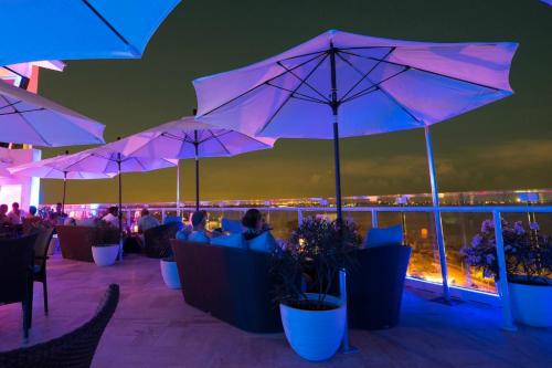 A rooftop lounge with umbrellas and seating, illuminated by colorful lights, overlooking a city skyline at night.